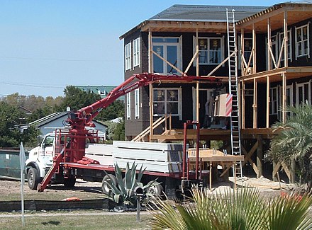 Drywall is delivered to a building site on a flatbed truck and unloaded with a forked material handler crane. The bulk drywall sheets are unloaded directly to upper floors through an open window or exterior doorway.