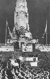 Foto en blanco y negro.  Ceremonia oficial con una multitud frente a una escalera que conduce a un alto monumento en forma de torre.