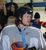 Een blanke hockeyspeler vanaf de borst omhoog.  Hij draagt ​​een grijze trui met een gestileerde bruine thrasher met een hockeystick als logo en een zwarte helm met vizier