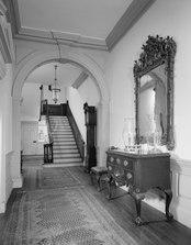 Un couloir avec des tapis s'étendant à travers une arche et menant à des escaliers au bout.  Une console avec des tiroirs se trouve à droite du hall avec un miroir au-dessus sur le mur.