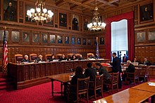 A room with ornate brown wooden paneling and oil portraits on the walls. At the left seven people wearing black robes sit behind a similarly decorated wooden bench, elevated slightly from the red-carpeted floor. On the right are several people in suits sitting at chairs behind tables. In the rear is a large window with red drapes.