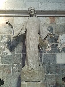 Statue en pierre blanche du Christ étendant les bras et vêtu d'une longue toge. Autour de son cœur sont dessinés des flammes et des rayons lumineux.