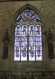 Au centre de l'image, vitrail légèrement bleutée représentant quatre femmes en robe. De part et d'autre du vitrail, des niches sont incluses dans un mur de pierre. Dans l'une des niches, une statue d'homme barbue est conservée.