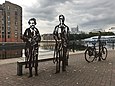 In einer Hafengegend stehen vor einer schmalen Bank zwei Stahl-Statuen. Die eine stellt eine Frau im langen Mantel dar, die ein Buch hält. Links von ihr ist ein Brillenträger im Anzug und Spazierstock. Abseits der beiden sind zwei weitere Statuen in Form eines Fahrrads und eines Vogels zu sehen.
