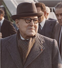 Photographie d'un homme souriant portant un manteau, des lunettes, un chapeau et une fine moustache.