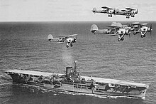 Photo en noir et blanc d'un porte-avions en mer, survolé par plusieurs biplans.