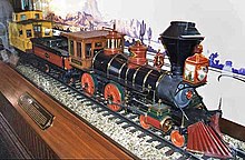 A ridable miniature steam locomotive, tender, and yellow-colored caboose in a display case with an image of a desert behind them