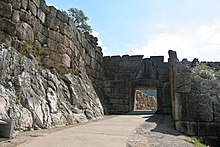 La entrada al patio que conduce a la Puerta del León.  A la izquierda, la pared rocosa está coronada por un muro de grandes piedras.  A la derecha, las ruinas de un bastión.  En el centro, el camino que conduce a la puerta propiamente dicha, una gran abertura coronada por una escultura triangular que representa a dos leones gateando uno frente al otro a cada lado de una columna, sus patas delanteras descansando sobre un altar.