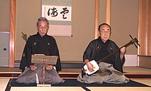 A Japanese man playing a shamisen while another sings Man playing shamisen.jpg