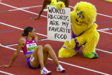 Photo of Sydney McLaughlin-Levrone sitting on red athletics tracks next to a furry yellow mascot holding a sign with the text "world records are my favorite food"