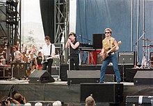 U2 playing on an outdoor stage. The Edge is on the left playing guitar, Bono in the center with a microphone, and Adam Clayton on the right playing bass guitar. A drum set is partially visible on the right side.