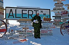 United States Ambassador David Jacobson in front of Alert's welcome sign which features many fingerposts pointing to places worldwide. The sign was erected prior to division and has NWT for the Northwest Territories. US Ambassador visits Alert, Nunavut.jpg