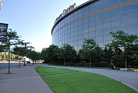 The wide, convex facade of a glassy sports arena and the sidewalk, lawn, and lampposts in front of it. The words "Xcel Energy Center" sit in red atop the arena.