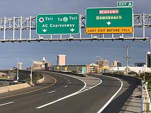 A three-lane freeway at an exit junction, with two green highway directional signs in the foreground and a skyline of hotels in the background.