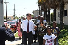 Un uomo bianco in camicia e cravatta, Eric Garcetti, cammina su un marciapiede accanto a un gruppo di neri di tutte le età, incluso un uomo che gli parla.