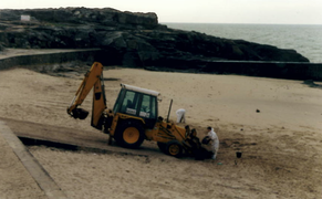 Photographie d’une pelleteuse orange s’avançant sur une plage, avec deux hommes en blanc.