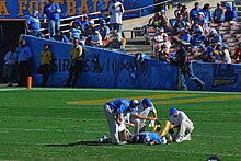 Foto de Jones na UCLA deitado em um campo cercado por treinadores de equipe