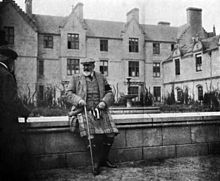 Édouard VII portant un kilt et une canne est assis sur un muret devant une maison à trois étages.