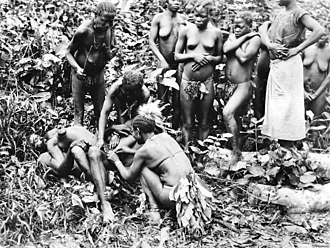Photographie d'une jeune femme nue, apparemment inconsciente, allongée sur le dos sur un homme dans la jungle. Deux femmes lui touchent l'aine sous le regard d'un groupe de femmes portant des bpagnes