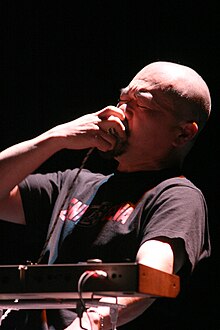 Der kahl rasierte Musiker hält sich, mit geschlossenen Augen, ein Mikrofon vor den Mund. Eine Kante der Rückseite des Synthesizers ist im Vordergrund zu sehen.