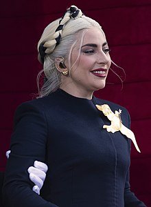 Side view of a smiling Lady Gaga, as she is looking away from the camera, wearing a navy blue dress decorated with a golden bird.