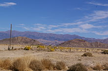 Sagebrush and tumbleweed lined desert with two mountain ranges visible in the distance. In the middle of this emptiness is a solitary sign post with a red, white, and blue sign.