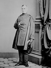 Full length photograph of a Civil War-era United States army officer. He is posed in a studio in front of a decorative column with one hand inside his coat.