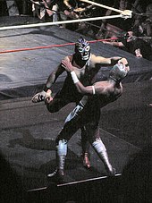 Foto eines maskierten Wrestlers, der einem anderen maskierten Wrestler eine Manschette gibt, während er seinen Kopf hält.