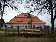 V pozadí stojí farní budova s červenou mansardovu střechou, vepředu oplocená socha svatého Jana Nepomuckého bez koruny, před plotem lavička