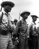 Tres hombres, vestidos con uniformes y sombreros, están de pie y miran hacia la derecha.  Están armados con granadas, pistolas y tienen valijas.  Se pueden ver otros hombres al fondo.