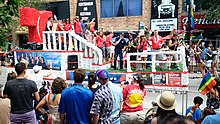 Несколько человек на параде проходят по улице, заполненной людьми.  В задней части поплавка, слева от изображения, находится большая красная буква «Т», представляющая логотип сети.  Несколько баннеров, рекламирующих программы Telemundo Chicago и инициативы по разнообразию, прикреплены к юбке платформы.