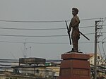 Side on view of a cement statue depicting a man with a moustache holding a rifle in one hand and a stick in another, wearing traditional clothes. He stands on a polished stone pedestal, and a city landscape and buildings can be seen in the background.