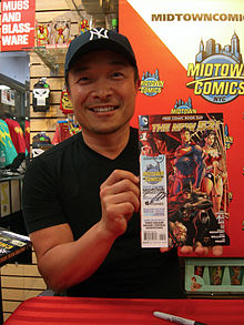 A photograph of a man wearing a black baseball cap and a black t-shirt looking at the viewer and smiling while holding a signed comic book