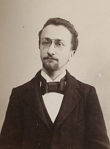 Photographie en noir et blanc d'un homme en costume complet portant une courte barbe et des lunettes.