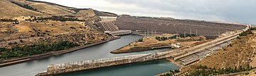 Long barrage vu d'en bas avec 8 grandes canalisations à gauche et déversoir en pente douce à droite : dans un paysage marron avec des bosquets d'arbres vert foncé.