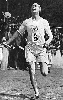 Photographie d'un homme en pleine course olympique, un seul pied au sol, tenant dans sa main droite un bâton de relais.