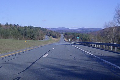 A four-lane highway in the woods looking toward mountains on a sunny day.
