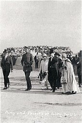 En uniforme militaire et portant un képi, le roi et la reine vêtue d'une longue tenue claire sont accompagnés par quelques personnalités officielles. Au loin, derrière eux, on distingue par une foule massée en un même endroit