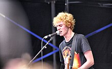 Byrne is singing into a microphone on its stand. He has a guitar strap but the instrument is out of shot. Behind him are blue beams from lights. He wears a David Bowie/Aladdin Sane t-shirt. He has curly gold-red hair with his eyes closed and veins popping in his neck.