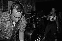 Foreground: Sweaty, make guitarist, and female singer, live on stage. In the background we see the bassist in the shadows, and hands of the drummer.