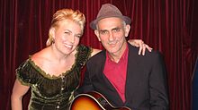 Upper body shot of a woman with Kelly. Both are smiling; she has her left arm across his back to his left shoulder. Kelly is wearing a hat, black jacket, and red shirt and is cradling a guitar (mostly out of view).