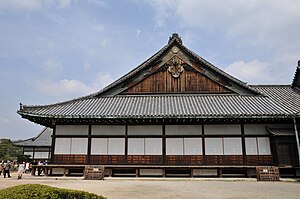Wooden building with a large hip-and-gable roof and white walls. The gable is decorated with a chrysanthemum symbol.