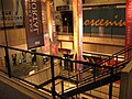 A multi-story interior atrium, made of concrete and with a staircase running downwards in the middle. Several multi-colored banners hang next to the stairs. The back wall has text slightly blocked from view by the banners, with the visible portion reading "osceniu" in cursive.