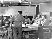 Nine men sit around a large table. Another is standing, leaning over the table. On the wall behind them are maps of the Pacific Ocean and Enewetak Atoll.