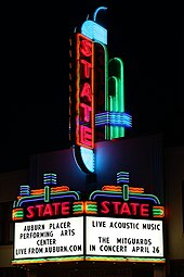 Photograph of a large, elaborate neon sign at night. The word "STATE" is written vertically in red neon tubing on a tower above a marquee. The marquee sign proper below the tower also has an elaborate neon tubing design, including the word "STATE" written horizontally in red neon tubing above each of the two panels facing the camera. A reader board on the front-facing panel has black lettering that says "AUBURN PLACER/PERFORMING ARTS/CENTER/LIVE FROM AUBURN.COM". A second reader board on a side panel says "LIVE ACOUSTIC MUSIC//THE MITGARDS/IN CONCERT APRIL 26".