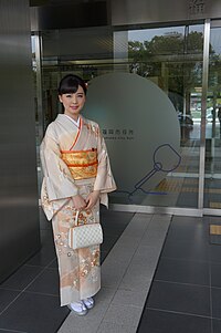 A woman standing outside a building wearing a short sleeved light pink kimono with a gold belt.