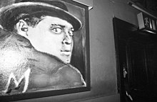 Photographie en noir et blanc montrant une peinture accrochée au mur, où figure un homme au regard halluciné avec la lettre M dans son dos.