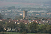 Roofs of several houses with the square tower of the church prominent amongst them. In the background are fields and hills.