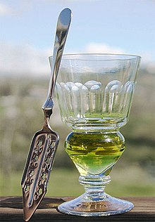 stemmed reservoir glass containing a green coloured liquid and a flat, slit, absinthe spoon
