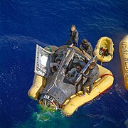 Fotografía en color de Armstrong y Scott en su cápsula esperando con el equipo de rescate la llegada de su barco.